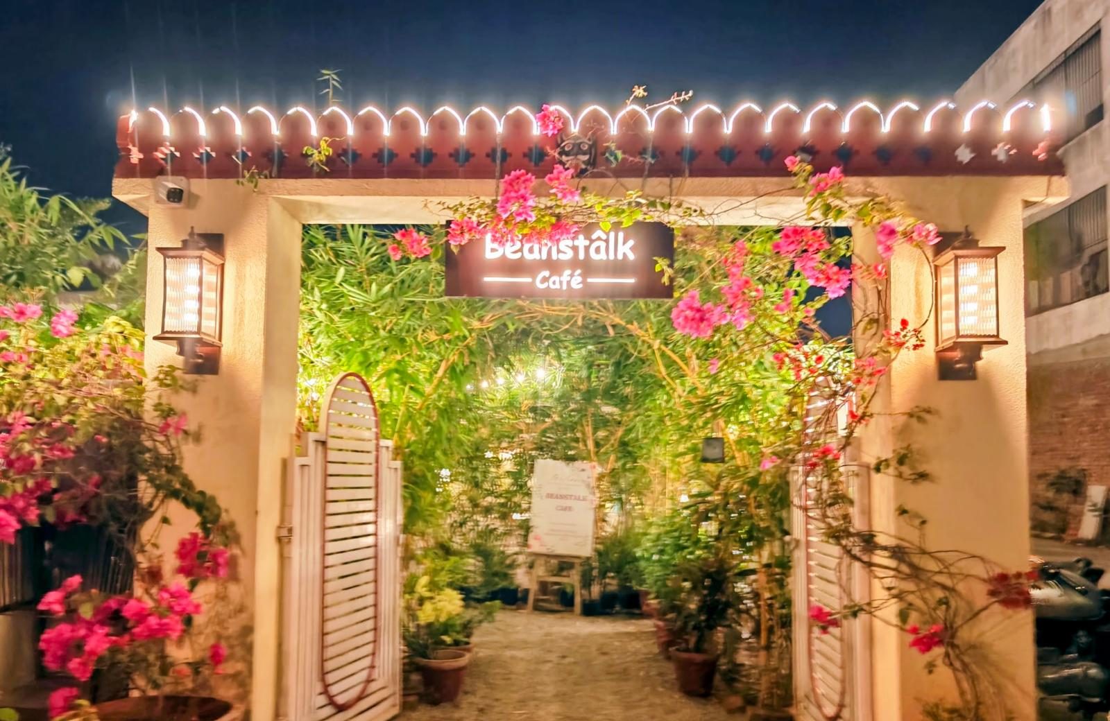 The Beanstalk Café gate at night: a lit sign under a scalloped Rajasthani parapet strung with lights, bougainvillea and open white shutter gates.